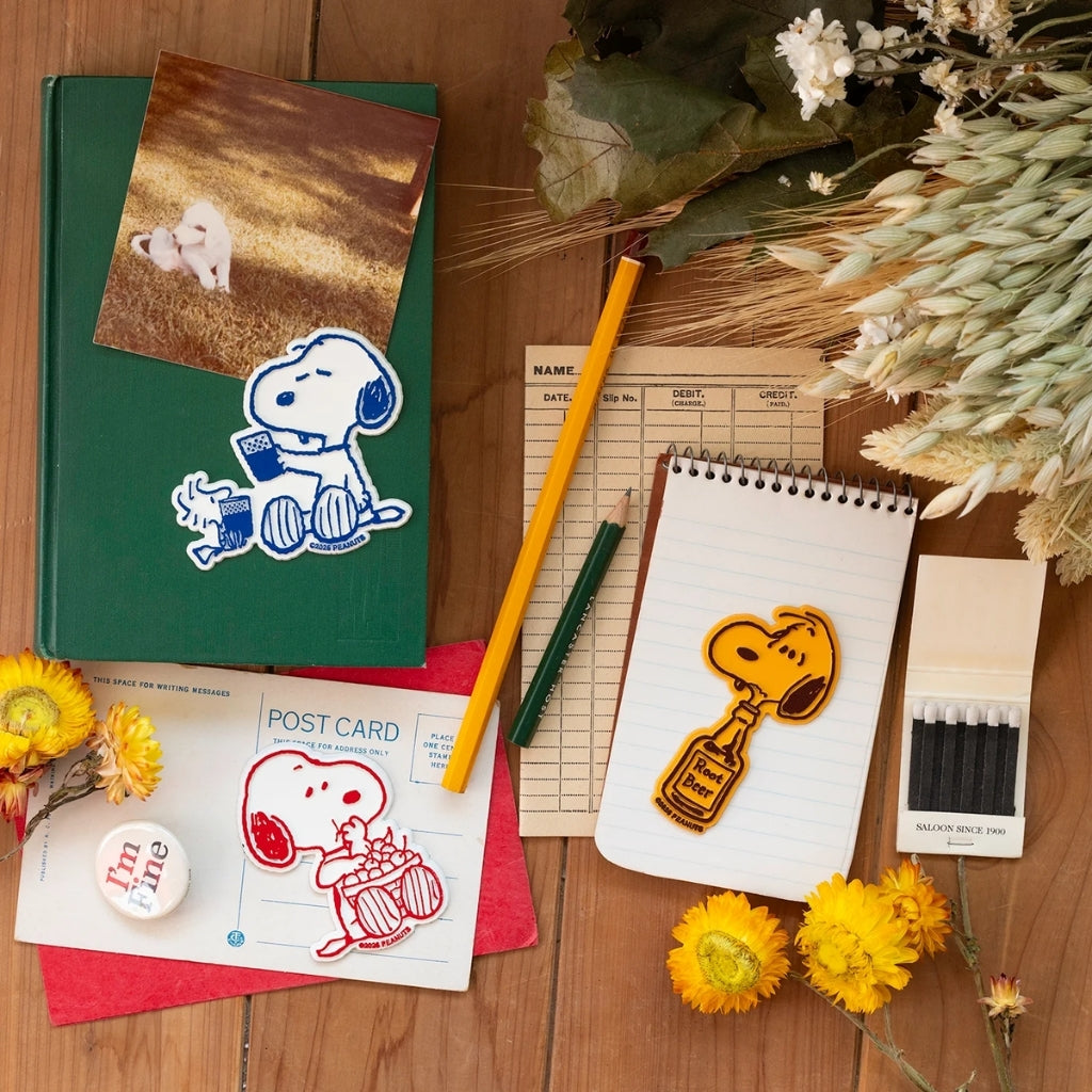 Snoopy Root Beer fridge magnet on a notepad, styled with other Peanuts Snoopy magnets, a vintage postcard, and dried flowers